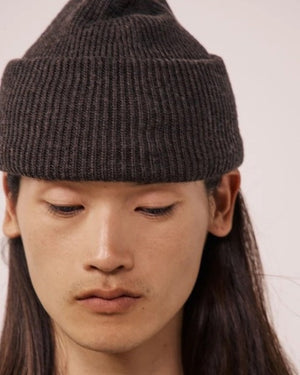 A person with long dark hair wears the HARTFORD - SEAMLESS WOOL BEANIE IN BROWN and a dark shirt, looking directly at the camera against a white background. The 100% wool beanie from HARTFORD adds both warmth and style to their look.