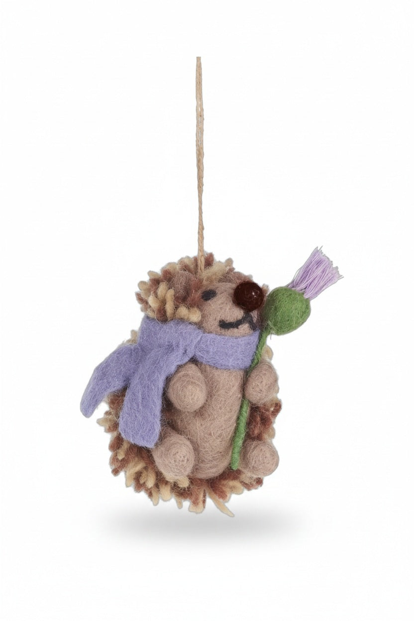 A THEA HEDGEHOG WOODLAND ORNAMENT by FELT SO GOOD, designed as a felt Scottish thistle hedgehog wearing a purple scarf and holding a flower, hanging from a twine loop.