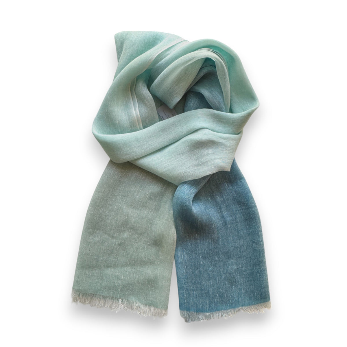 The BAJRA - HAND DYED LINEN BLEND SCARF IN MEMORY OMBRE offers a soft, lightweight feel with a mint to teal gradient and traditional Nepal-inspired design. Its pashmina-like fabric is slightly sheer, ending in fringed edges for an elegant drape on any neutral background.