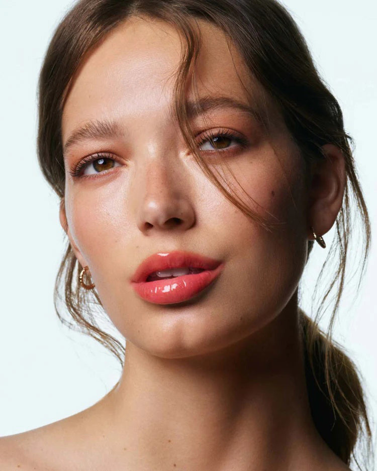 A woman with light skin and brown hair tied back wears small gold hoops and soft peach makeup. Her glossy pink lips, thanks to U BEAUTY’s THE PLASMA LIP COMPOUND IN BELLINI, catch the light as she gazes softly at the camera, a strand of hair falling across her face.