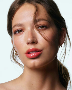 A woman with light skin and brown hair tied back wears small gold hoops and soft peach makeup. Her glossy pink lips, thanks to U BEAUTY’s THE PLASMA LIP COMPOUND IN BELLINI, catch the light as she gazes softly at the camera, a strand of hair falling across her face.