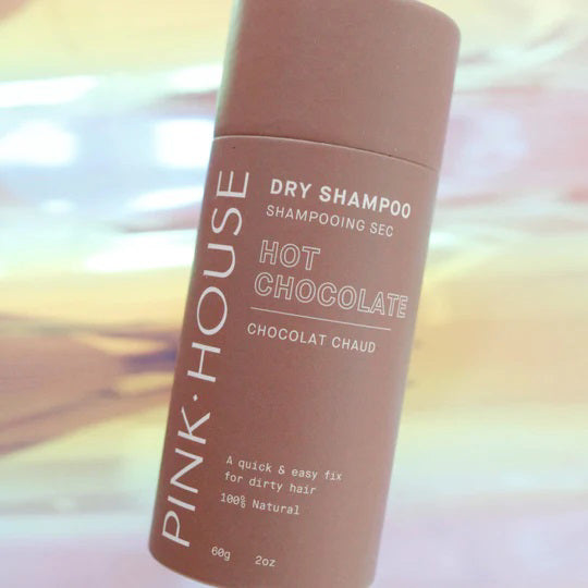 A cylindrical bottle of PINK HOUSE ORGANICS - DRY SHAMPOO HOT CHOCOLATE by PINK HOUSE ORGANICS is shown. The label highlights its 100% natural ingredients and promotes it as a quick and easy solution for greasy hair. The bottle is displayed against a pastel-colored, blurred background.