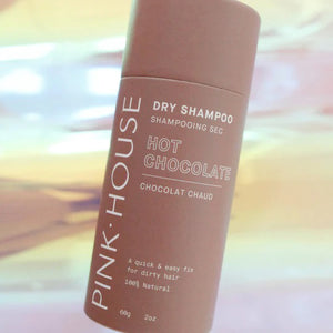 A cylindrical bottle of PINK HOUSE ORGANICS - DRY SHAMPOO HOT CHOCOLATE by PINK HOUSE ORGANICS is shown. The label highlights its 100% natural ingredients and promotes it as a quick and easy solution for greasy hair. The bottle is displayed against a pastel-colored, blurred background.
