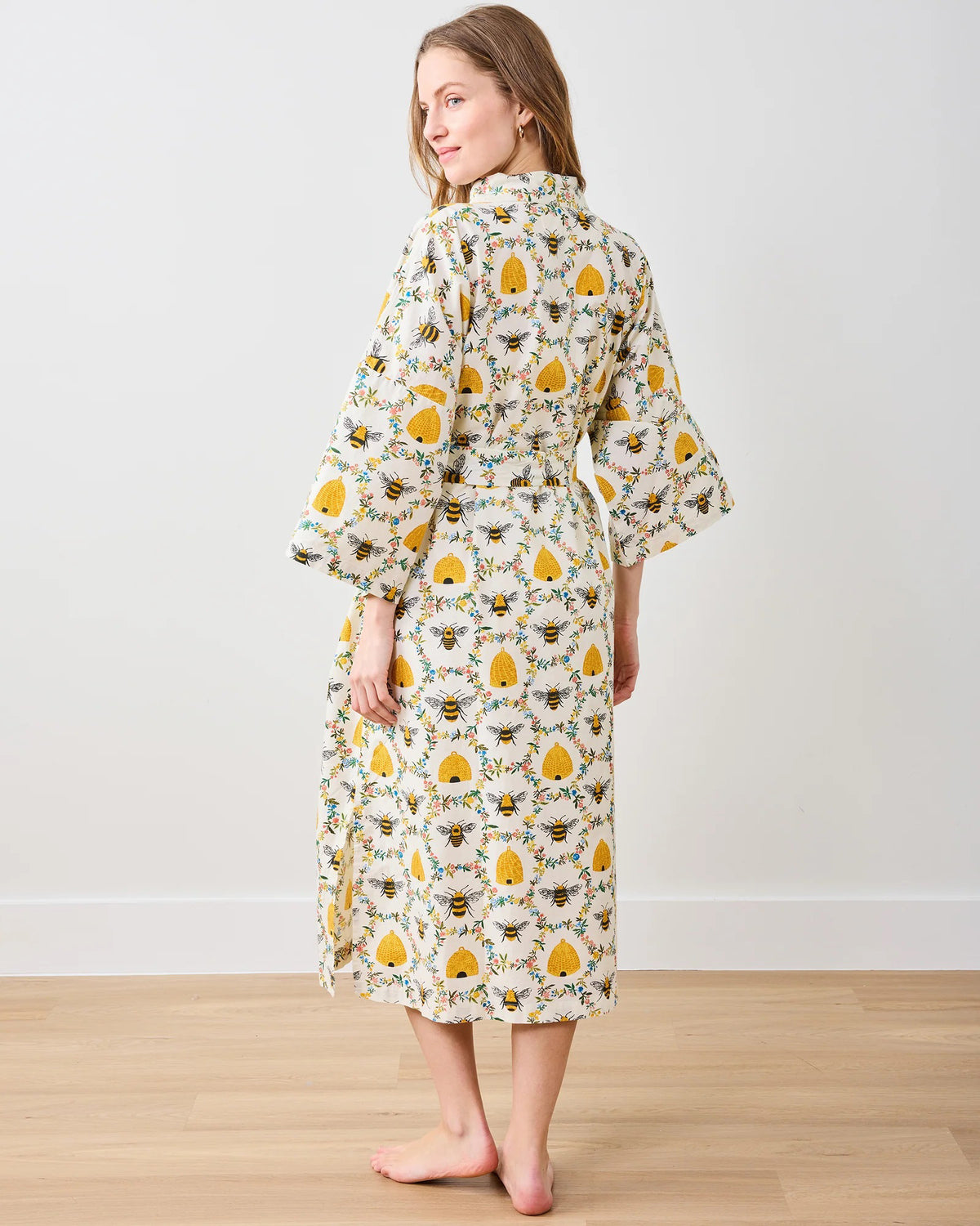 A person models the PRINTFRESH - SWEET BEE COTTON ROBE, featuring a floral design with sunflowers and leaves. Made from organic cotton, it has long sleeves and a waist tie, set against a plain white wall.