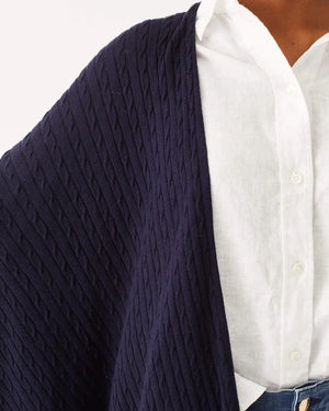 A woman with short, curly hair smiles while wearing a white blouse, wide-leg blue jeans, and the MERSEA CHARLESTON CABLE WRAP over her outfit, standing against a plain white background.