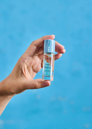 A LAFCO FINE FRAGRANCE, TROPIC WATERS 10ML box stands upright in a halved coconut, water splashing around it against a neutral background—an evocative display for this long-lasting scent inspired by the essence of coconut water.
