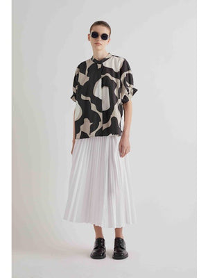 A person with brown hair in a low bun is shown from the back, wearing the FRANK AND EILEEN - MOONBEAM TOP by MEIMEIJ—an abstract print top with loose, short sleeves in black, white, and beige—paired over a white pleated skirt.