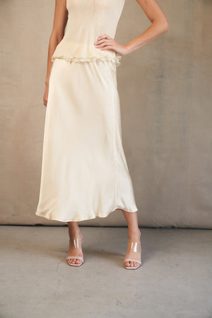 A woman models the ROSE & CROWN - SATIN MIDI SKIRT in silky blue, paired with a short-sleeved light blue knit top, standing against a white background. Her head is not visible in the image.