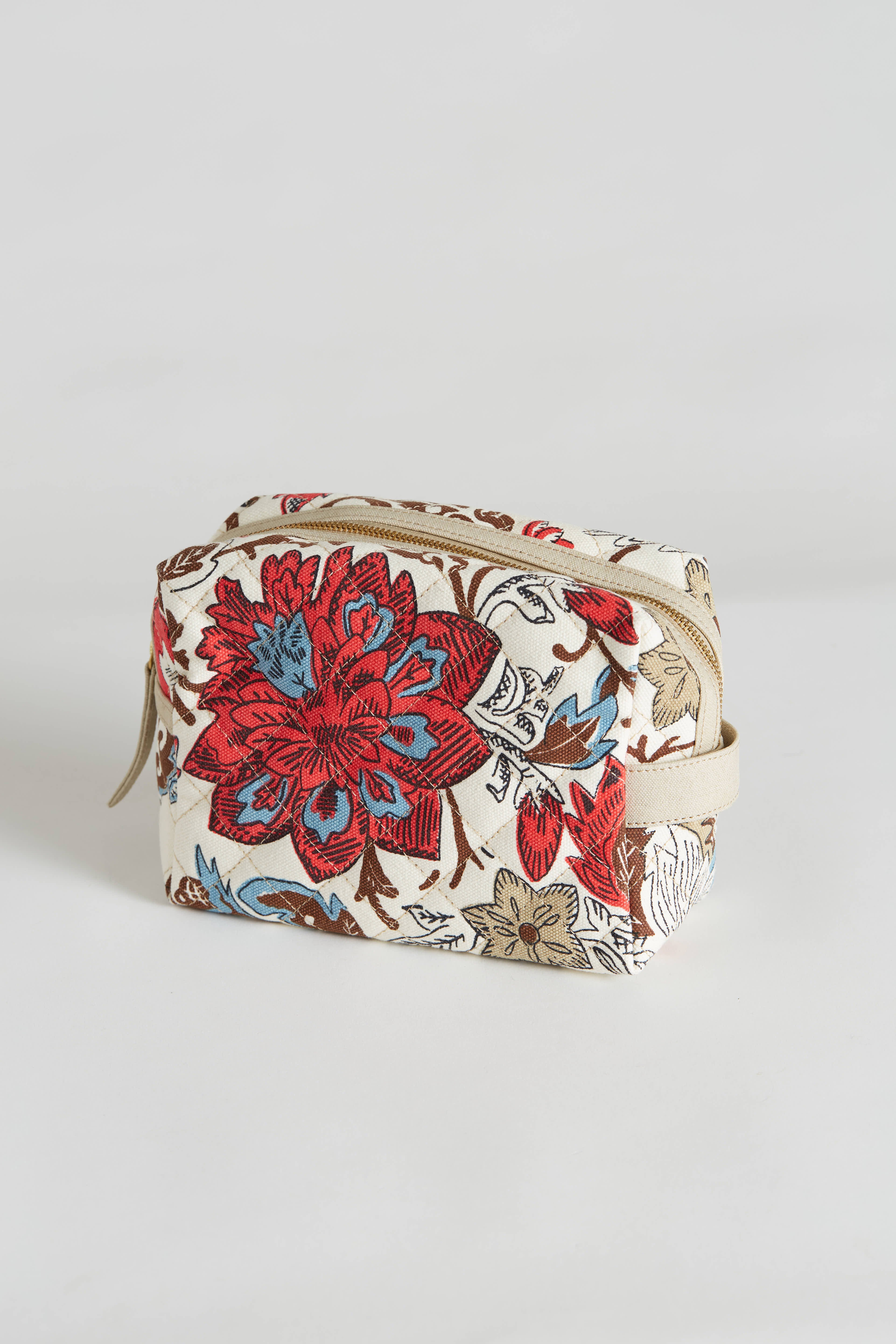 The INOUI EDITIONS - PIPPA COSMETIC BAG by INOUI is a small, rectangular, quilted cotton cosmetic bag with a beige base. It features a vibrant floral pattern in red, blue, and brown colors and measures 21x10 cm. The bag has a zipper closure on top and includes a polyester lining. It is displayed against a plain white background.