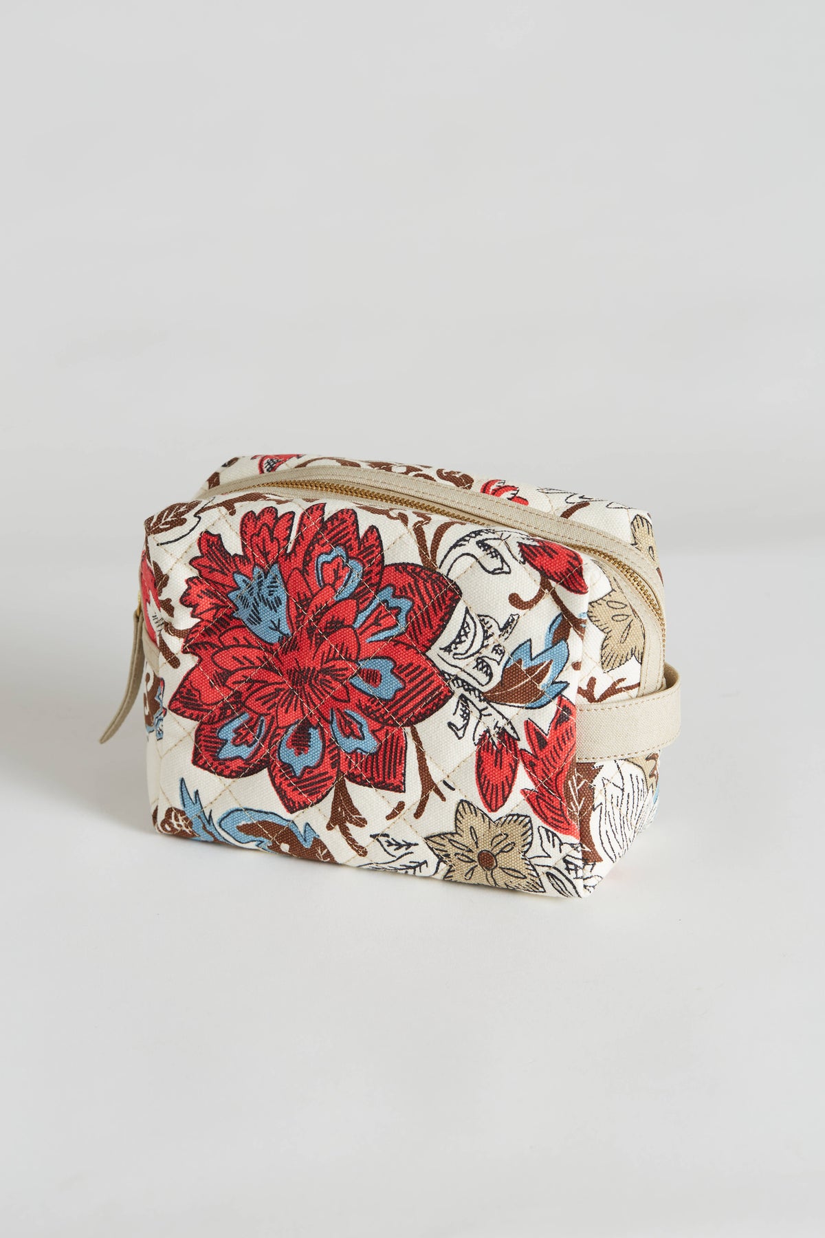 The INOUI EDITIONS - PIPPA COSMETIC BAG by INOUI is a small, rectangular, quilted cotton cosmetic bag with a beige base. It features a vibrant floral pattern in red, blue, and brown colors and measures 21x10 cm. The bag has a zipper closure on top and includes a polyester lining. It is displayed against a plain white background.
