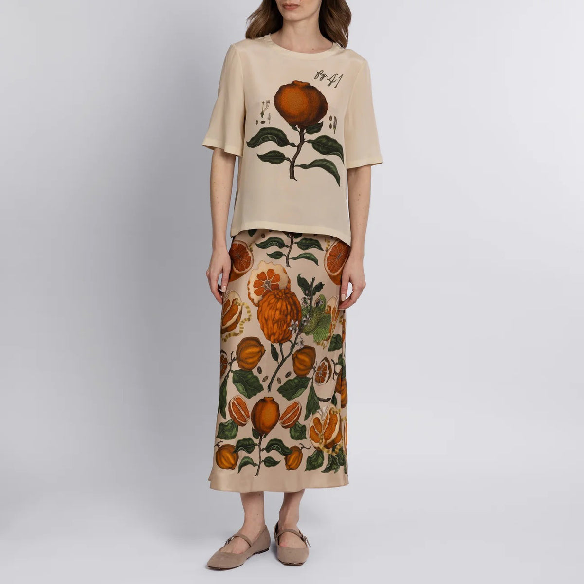 A person stands barefoot on a wooden floor wearing the SABINA SAVAGE - ORANGES AND LEMONS SILK BIAS SKIRT from SABINA SAVAGE ENGLAND, featuring an orange and green floral and fruit exotic print. The plain white background highlights the skirt&#39;s elegant bias cut.