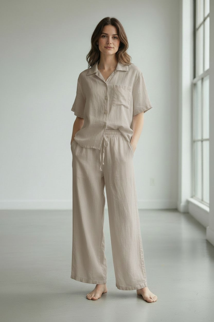 A person wearing the LLEA BLUE - DANA Straight Leg Drawstring Linen Pant in Oatmeal stands barefoot on a light gray floor in a minimal, bright room.