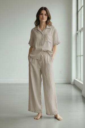 A person wearing the LLEA BLUE - DANA Straight Leg Drawstring Linen Pant in Oatmeal stands barefoot on a light gray floor in a minimal, bright room.