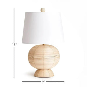 The NAPA HOME AND GARDEN Maye Rattan Hourglass Lamp with Shade features a wicker-style base and white shade, casting a warm glow on any wooden surface.