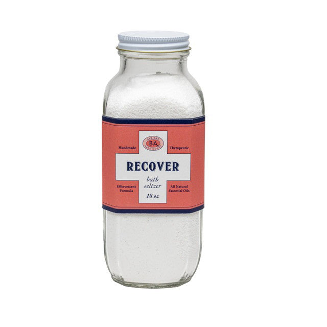 A clear glass bottle labeled &quot;BAUDELAIRE - RECOVER BATH SELTZER 18OZ&quot; with a red and white label. The label highlights that it is handmade, therapeutic, features an effervescent formula, contains all-natural essential oils, and weighs 18 ounces.