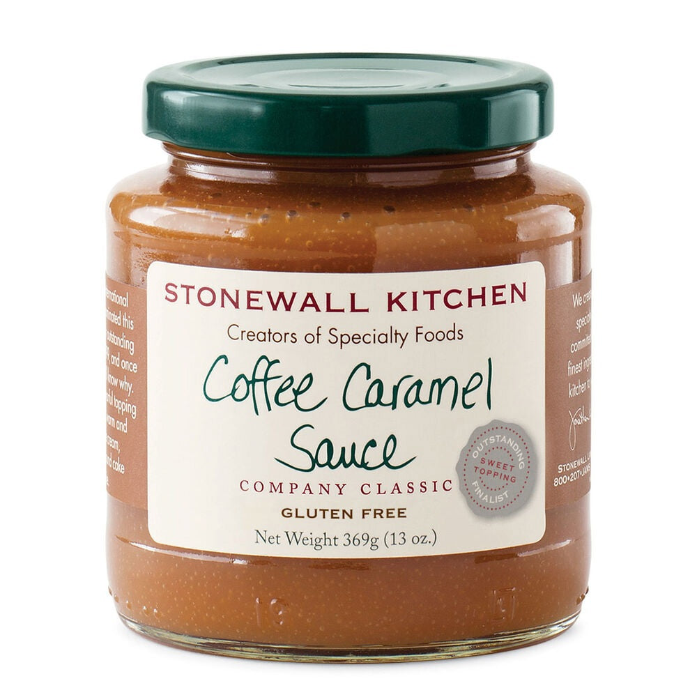 A jar of STONEWALL KITCHEN - COFFEE CARAMEL SAUCE is displayed alongside a stick of butter, brown sugar, coffee beans, a glass of syrup, a small glass of oil, and a metal cup of cream on a wooden surface. This decadent caramel sauce from STONEWALL KITCHEN provides a rich and buttery sweetness that’s perfect for elevating your favorite desserts.