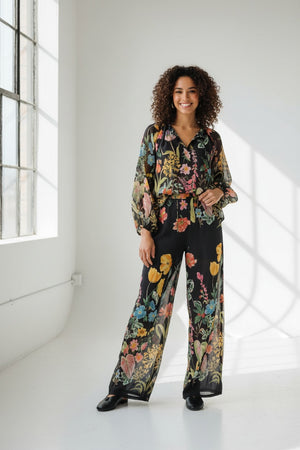 A person stands facing forward, wearing the LAVI - APHRODITE BOTANICAL BLOUSE by LAVI—a loose-fitting, long-sleeved statement blouse featuring a colorful floral pattern—against a plain white background.