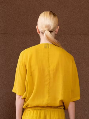 A woman with naturally curly hair models the NUDE - TIE WAIST V NECK BLOUSE by NUDE, a loose-fitting yellow top in a soft Tencel-cotton blend with short sleeves and an adjustable drawstring waist, against a plain light background.