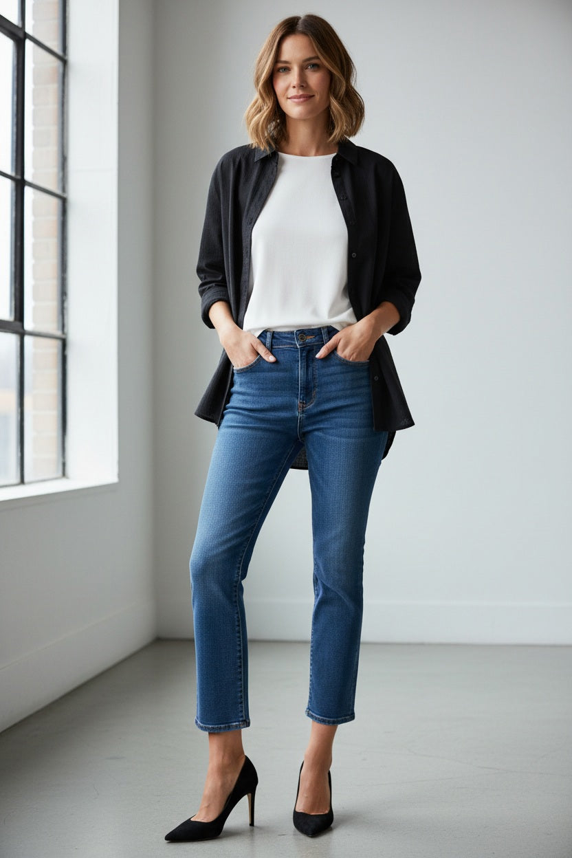 A woman with her hair pulled back stands against a plain background wearing SPANX Redefine Kick Flare Jeans in Medium Indigo, a black button-up shirt, black pointed-toe heels, and carries a black shoulder bag with one hand in her pocket.