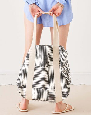 A woman in an orange long-sleeve shirt and denim shorts carries the MERSEA LEO TOTE BAG, a high-quality cotton tote from MERSEA with wide cream straps and an open top, featuring colorful orange, pink, and white checkered squares.
