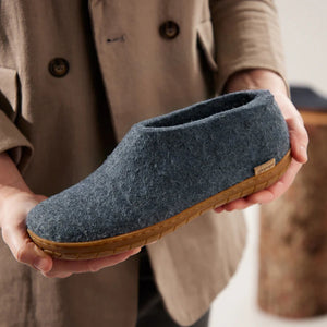 A person in a beige coat holds the GLERUPS SHOE WITH NATURAL RUBBER SOLE IN DENIM—cozy, itch-free slippers by GLERUPS, featuring gray natural wool and a brown rubber sole with a branded side label.