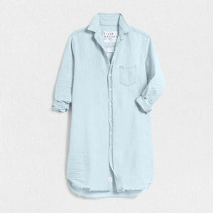 The FRANK AND EILEEN - MARY CLASSIC SHIRTDRESS in Classic Blue Tattered Wash by FRANK & EILEEN is a light blue, long linen shirtdress featuring a button-down front and long sleeves rolled up to three-quarter length. It has a single chest pocket and resembles the casual elegance of Famous Denim with its textured fabric and slightly frayed hem, displayed against a textured white background.