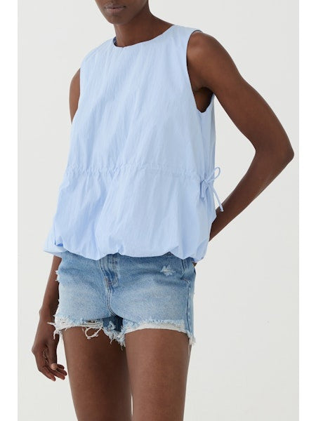 A person wears the ELEH - SLEEVELESS ROUND NECK TOP in light blue with a loose, blouson hem and drawstring waist, paired with light blue frayed denim shorts. The background is plain white.