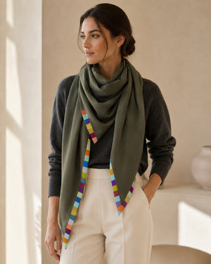 A woman stands indoors in soft natural light, wearing the FRANCO FERRARI Cashmere Scarf 135x135 in green—its colorful square trim elegantly draped over a dark gray sweater and light beige pants.