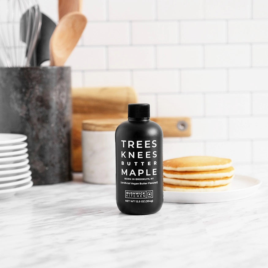 A bottle of "Trees Knees Butter Maple Syrup" by Bushwick Kitchen, originated in Brooklyn, NY. The label indicates it's artificially flavored vegan butter and gluten-free. The black bottle with white text has a capacity of 11.5 oz (326g) and features a black screw-on cap.
