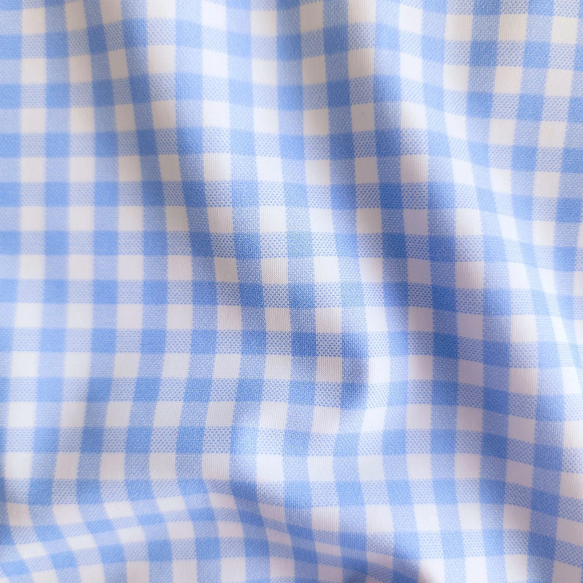 A man wearing COLLARS AND CO's Oxford Button Down Collar Apex Performance Dress Shirt in light blue gingham stands outdoors by a car, with green hedges and palm trees along a sunny street in the background.