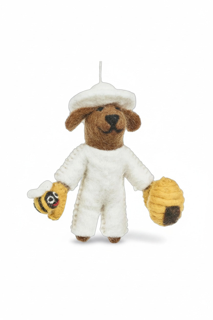BUMBLE THE BEEKEEPER ORNAMENT is a needle felted dog dressed in a white beekeeper suit, holding a small bee in one hand and a beehive in the other.