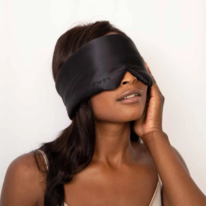 An older person with long gray hair wears a luxurious beige DROWSY - SATIN SLEEP MASK by DROWSY SLEEP CO, made of mulberry silk. In a matching satin top, they stand against a plain white background, ready for beauty sleep.