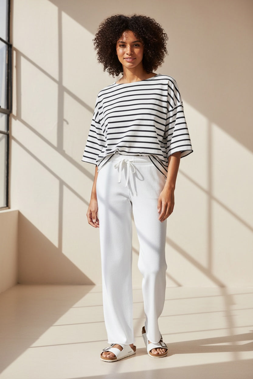 A person stands on a sandy beach facing the ocean, wearing FRANK &amp; EILEEN CATHERINE Full Length Favorite Sweatpants from the California Heritage Collection, a black and white striped top, and white sandals with bows.