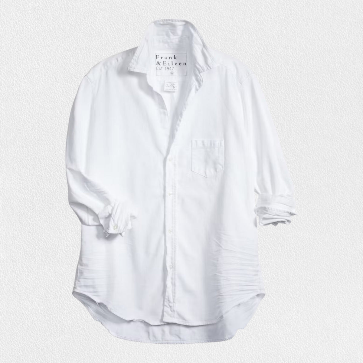 A woman with long brown hair stands in a relaxed fit pose, smiling and looking to the side. She wears the FRANK &amp; EILEEN - EILEEN RELAXED BUTTON UP IN DENIM WHITE TATTERED WASH and famous denim dark pants. Behind her is a stone porch with a potted plant and a wooden door, creating a cozy backyard setting.