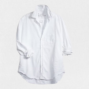 A woman with long brown hair stands in a relaxed fit pose, smiling and looking to the side. She wears the FRANK & EILEEN - EILEEN RELAXED BUTTON UP IN DENIM WHITE TATTERED WASH and famous denim dark pants. Behind her is a stone porch with a potted plant and a wooden door, creating a cozy backyard setting.