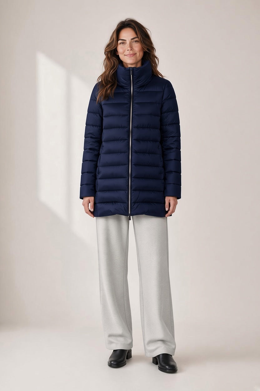 A woman wears a knee-length, dark blue SAVE THE DUCK - LYDIA PUFFER COAT IN NIGHT BLUE made of recycled nylon, featuring a high collar and a full-front zipper.