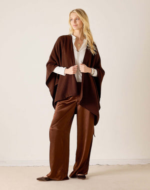 A woman with blonde hair stands by a sunlit window wearing the MERSEA - MODERN TRAVEL WRAP IN CHOCOLATE, featuring a modern bow detail. She pairs it with a black top and pants, accessorizing her look with bracelets and a necklace.