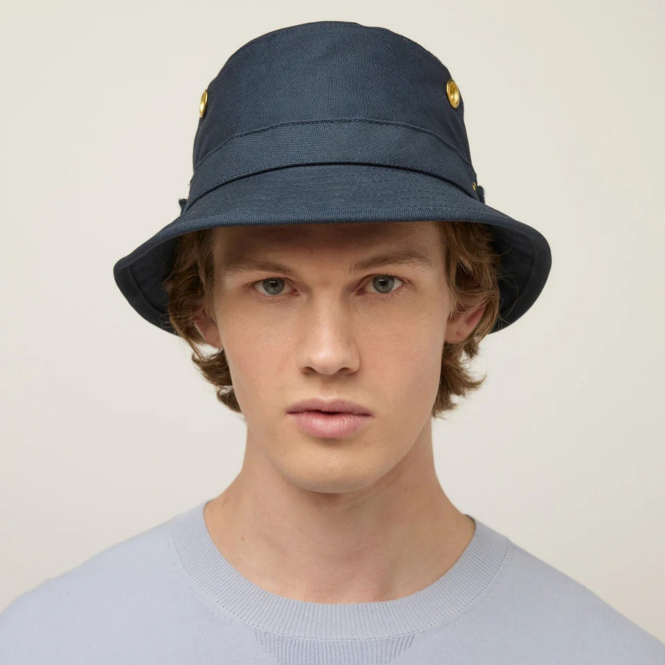 A young man faces forward against a plain background, wearing the TILLEY - ICONIC T1 BUCKET HAT in navy blue with gold side buttons and a light blue crew-neck shirt—a classic UPF 50+ sun protection style by TILLEY.