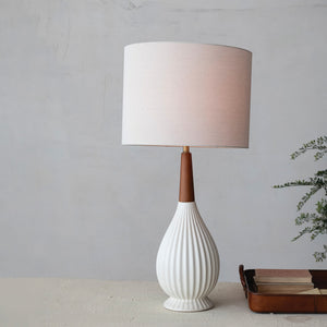 A cozy armchair with a black and white throw blanket sits by a marble side table holding wine. Nearby is the CREATIVE COOP PLEATED STONEWARE TABLE LAMP WITH RUBBERWOOD NECK AND LINEN SHADE, a potted plant, and a landscape painting on the wall.