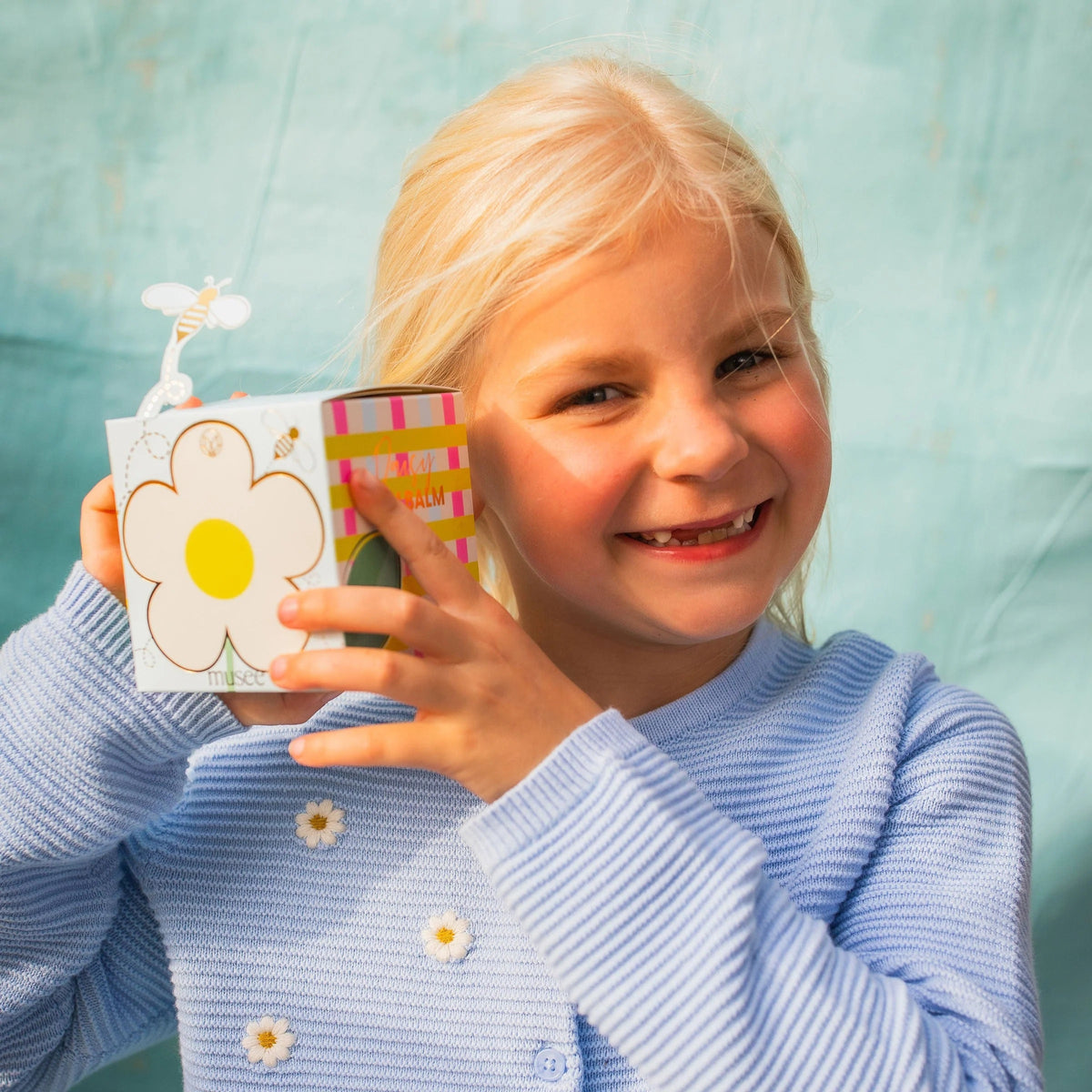 A light blue MUSEE - DAISY BATH BALM by MUSEE rests beside a beautifully decorated box featuring a white daisy with a yellow center and playful cartoon bees, including one bee attached on a wire. This delightful set is perfect for anyone who loves natural bath products and enjoys indulging in soothing bath balms.