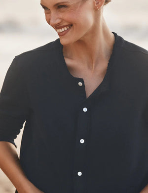 A woman with light brown hair stands outdoors by the beach, wearing FRANK & EILEEN’s JACKIE Sweatshirt Cardigan in British Royal Navy and blue jeans. She gazes into the distance with a relaxed expression; the background is softly blurred.