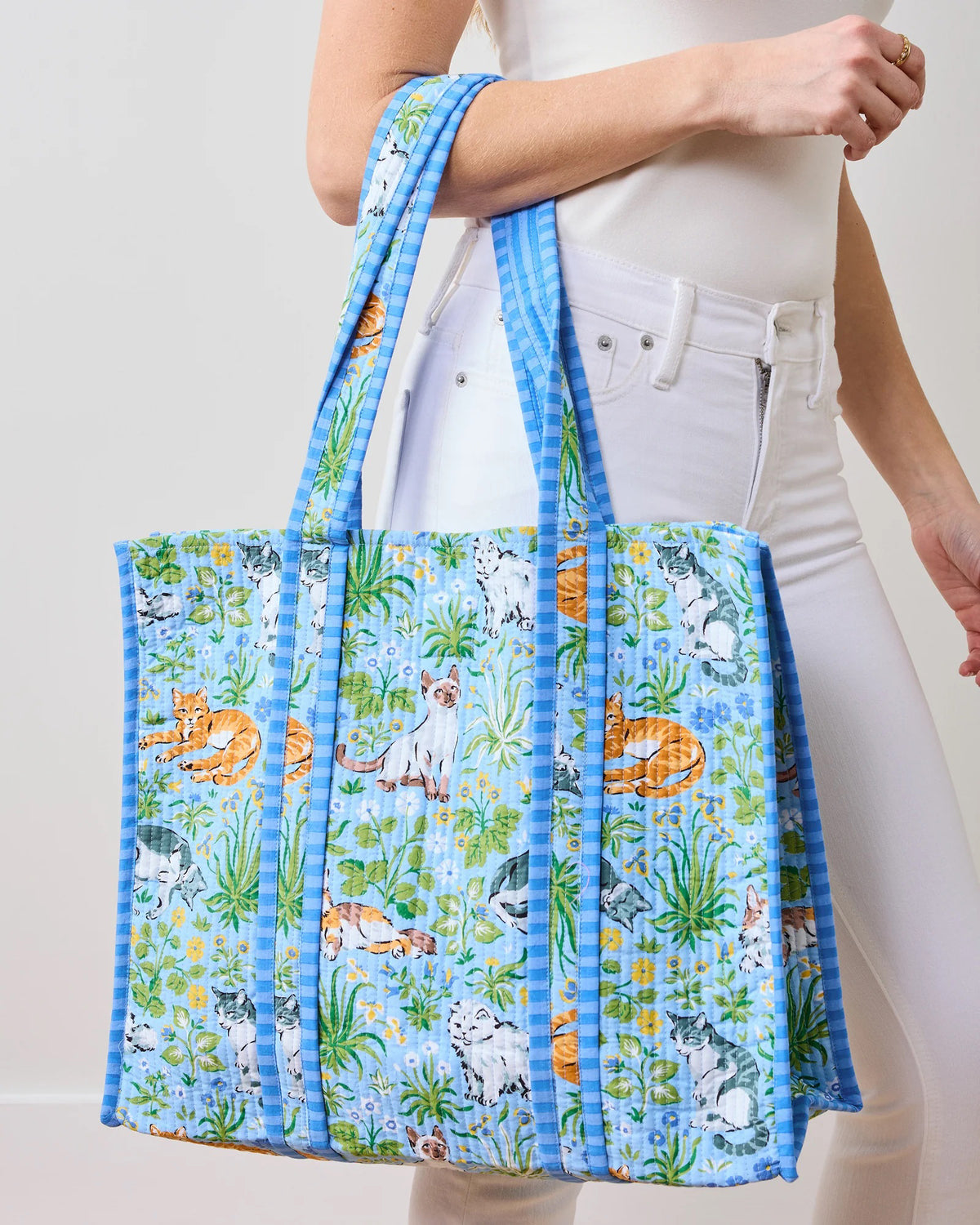 A person in white clothes holds the PRINTFRESH - MUST LOVE CATS TRAVELER TOTE, a light blue quilted cotton bag featuring cats among flowers and greenery. With its spacious design and long handles, it&#39;s ideal for any cat lover.