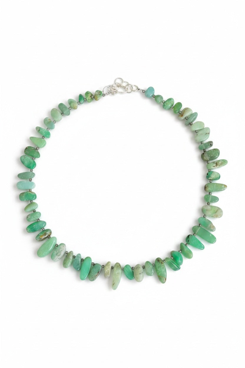 The ANN LIGHTFOOT Ras du Cou Necklace of Chrysoprase Pebbles features irregularly shaped green chrysoprase stones and a sterling silver clasp, elegantly displayed in a circular arrangement on a white background.