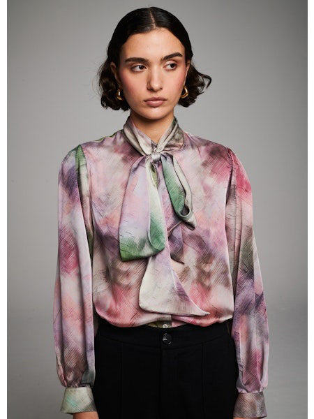 A woman with short dark hair wears the MILVA MI - BRUSHSTROKE BLOUSE by MILVA, a pastel long-sleeved statement bow top, paired with high-waisted black pants. She poses with eyes closed against a plain light background.