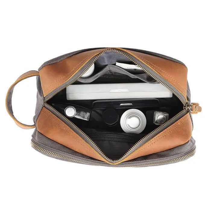 The TSD BRAND - Turtle Ridge Toiletry Bag in grey features waxed canvas, leather details, and a zip closure. It is open to show neatly organized travel essentials inside—bottles, a tube, a brush, and a white rectangular item.