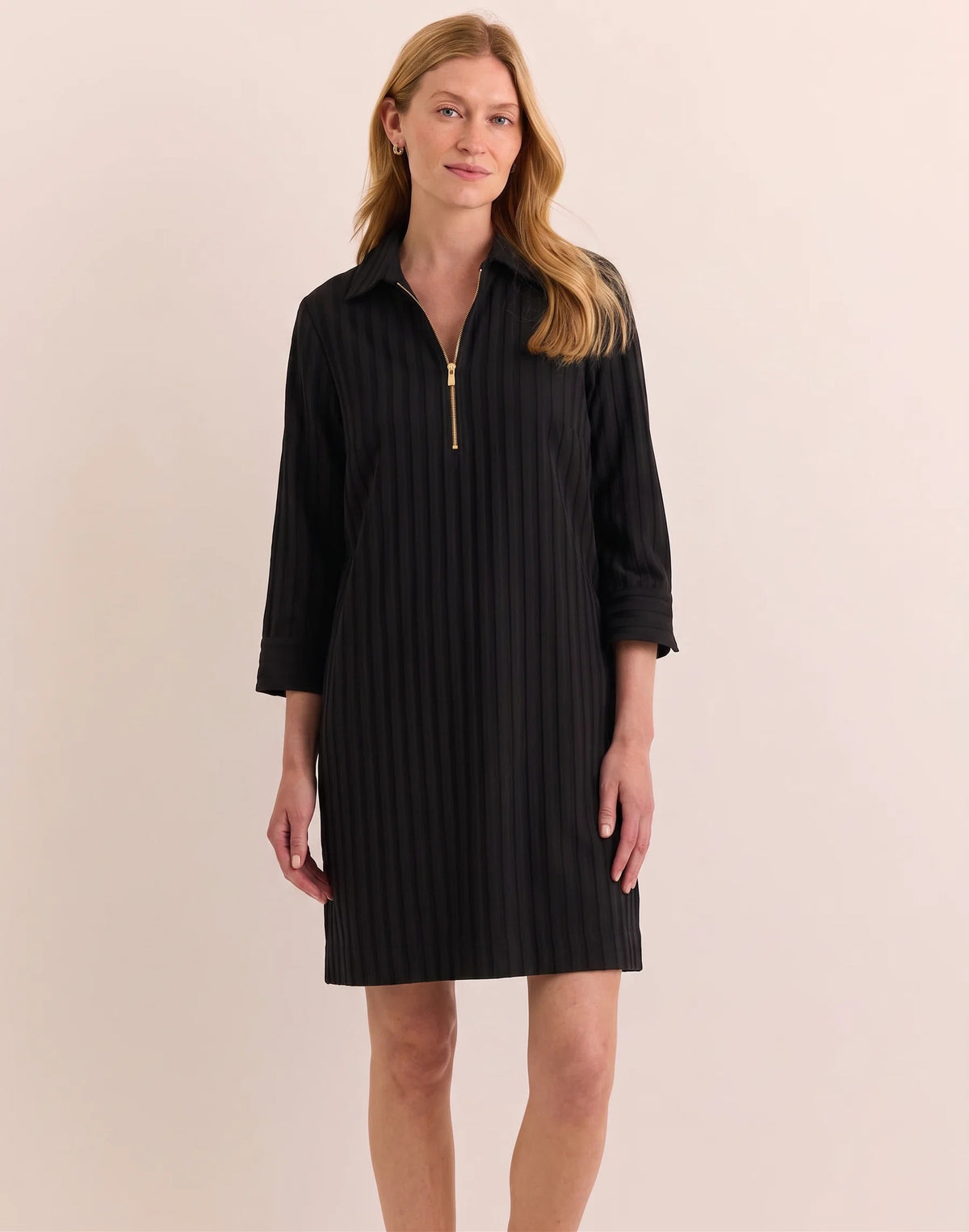 A woman with light brown hair and a slight smile stands against a light background, wearing the HINSON WU - Alessa 3/4 Sleeve Dress in Classic Stripe Black. This knee-length dress features vertical stripes, three-quarter sleeves, and a gold zip-up collar. Crafted from breathable fabric by HINSON WU, the look seamlessly blends style with comfort.
