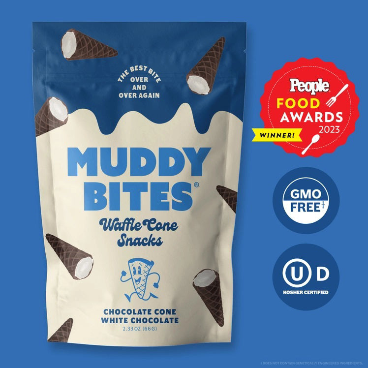 A person dressed in a light blue outfit sits on a plush, royal blue rug holding a bag of Muddy Bites Cookies &amp; Cream Waffle Cones in one hand and a small white chocolate waffle cone snack in the other. The beige and blue bag features an illustration of chocolate-dipped waffle cones.
