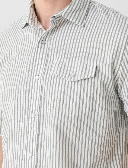 A young man, smiling with one hand in his pocket, wears CRIQUET’s Short Sleeve Performance Seersucker Pearl Snap Shirt in Sage and navy blue pants.