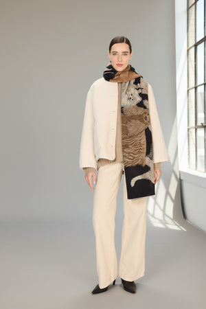 A woman stands facing forward against a plain white background, wearing a cream jacket, wide-leg cream pants, black shoes, and a brown top. Draped stylishly over her shoulders is the INOUI EDITIONS - TOUTOU DOGGIE WOOL SCARF by INOUI.