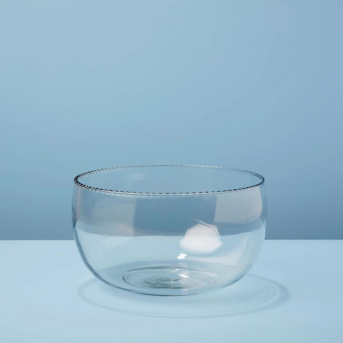 The RUFFLE GLASS BOWL LARGE from BE HOME, INC sits empty on a light blue surface against a matching background. This bowl, part of the Ruffle Glassware collection, boasts a decorative beaded rim and features a simple, modern design with unique texture that casts a subtle shadow.